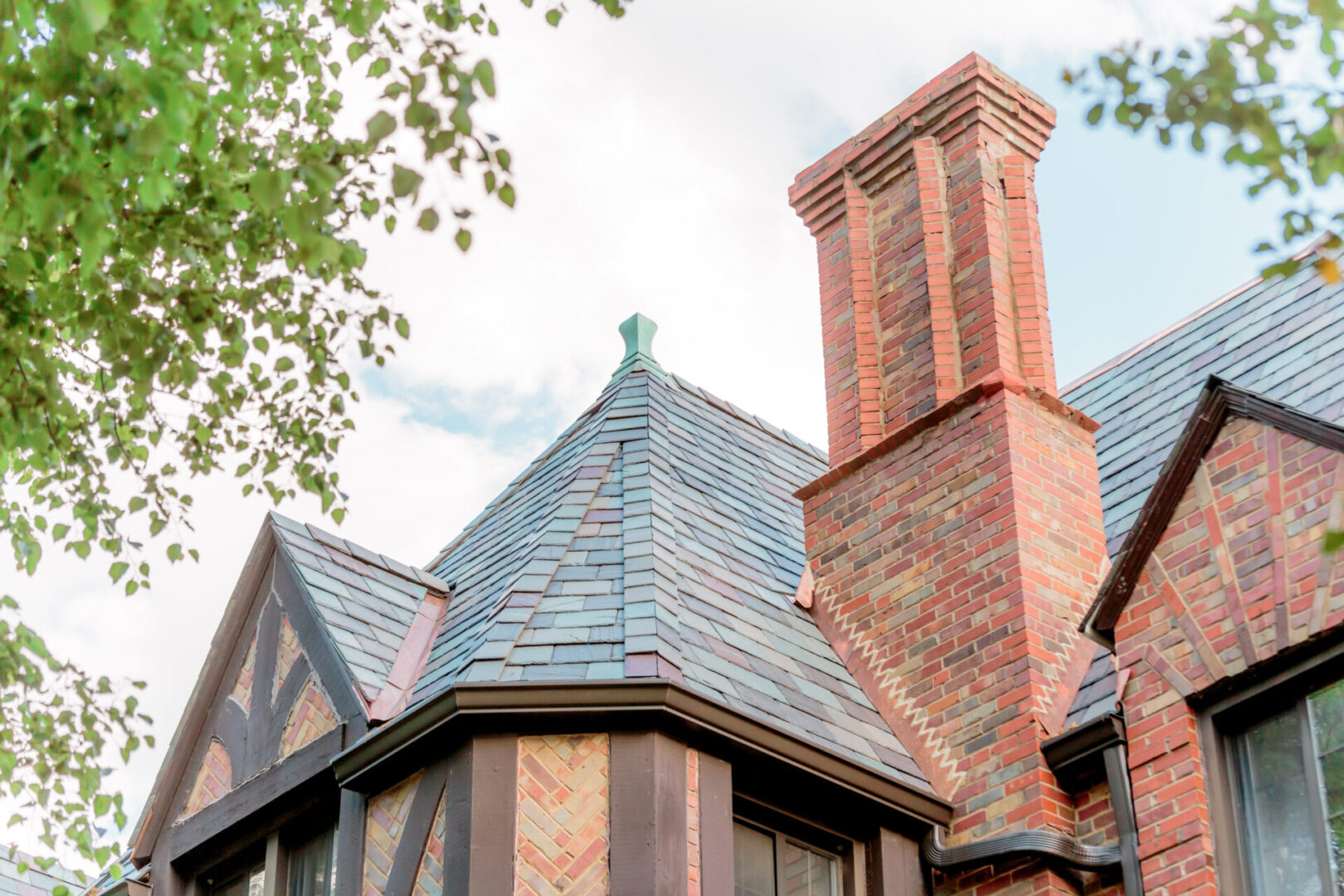 Architectural detail of a slate roof with a brick chimney under a cloudy sky.